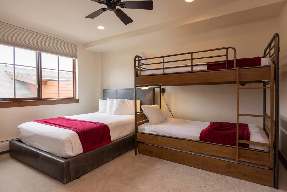 A family-friendly bedroom with a queen-sized bed, a bunk bed, red accent pillows, and a large window that fills the room with light. The space feels inviting and perfect for families or groups with children.