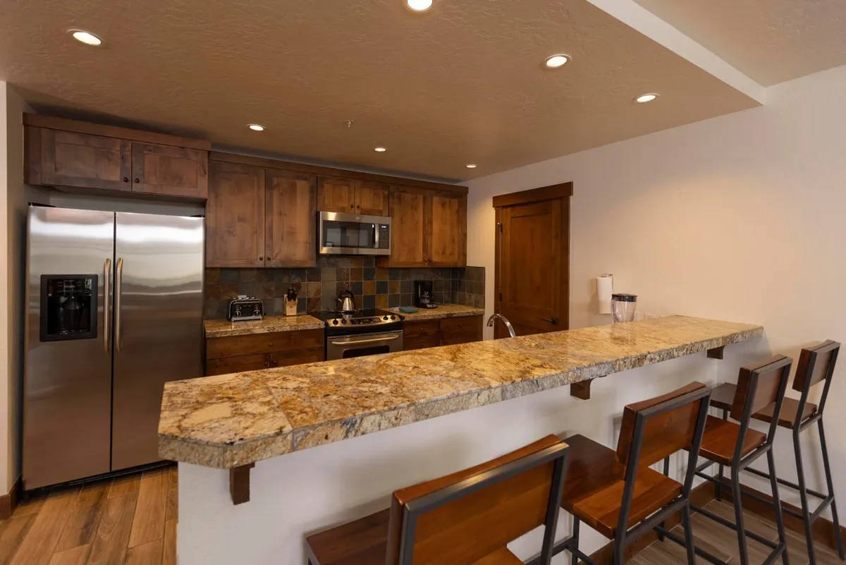 This image features a beautifully designed kitchen with gleaming granite countertops, rich wooden cabinetry, modern stainless steel appliances, and bar stools tucked along the counter. The open layout allows for visual connection to the living and dining areas, suggesting a space that fosters social interaction and culinary creativity.