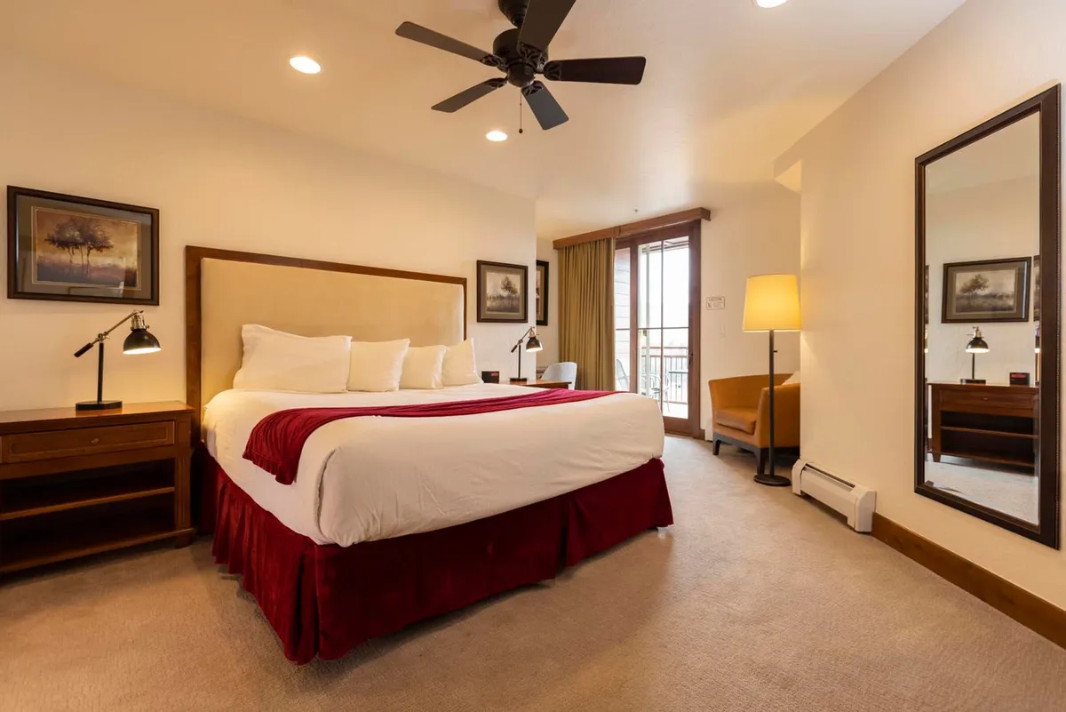 A tranquil bedroom featuring a plush bed with a red skirt, wooden furnishings, framed artwork, and a mirror reflecting the room’s soft natural light. The ambiance is peaceful, suggesting a restful retreat ideal for ending adventurous days.