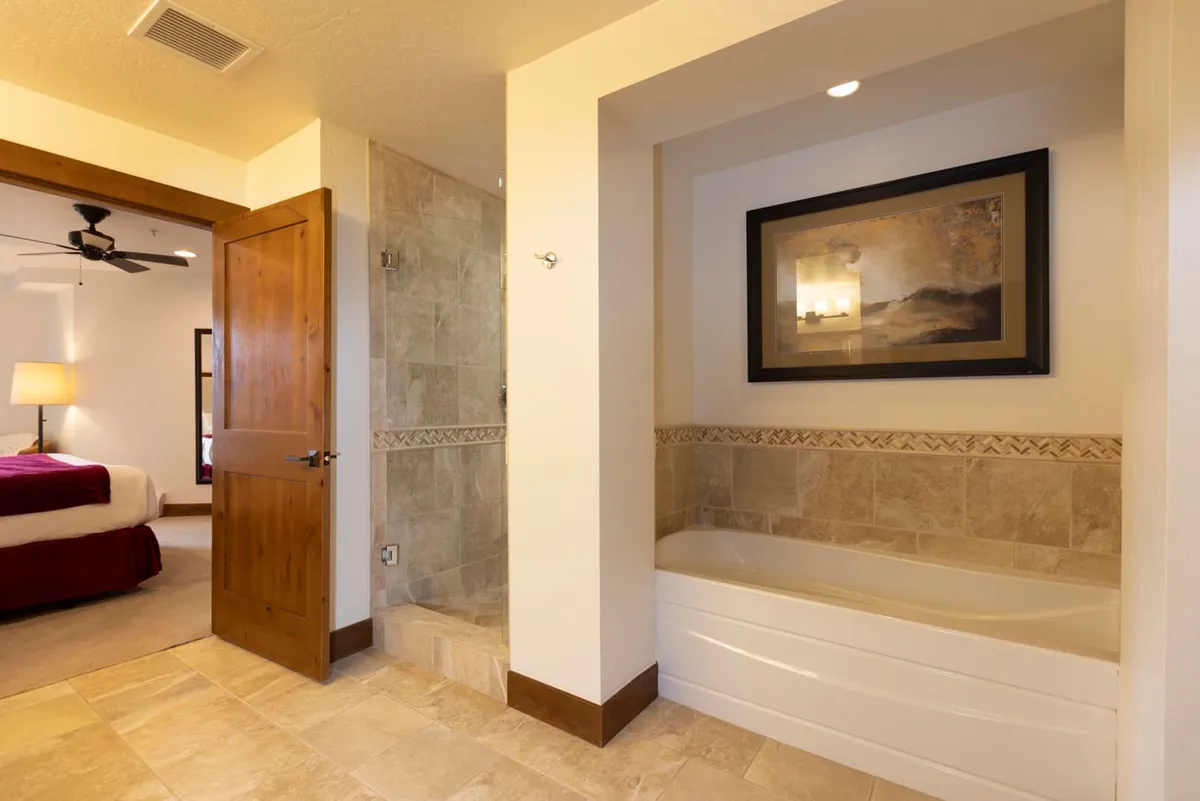 A serene interior shot of a luxurious bathroom, featuring a spacious bathtub, shower area, tasteful artwork, and a doorway leading to the bedroom. The space exudes tranquility and offers a glimpse of the spa-like experience available within the resort.