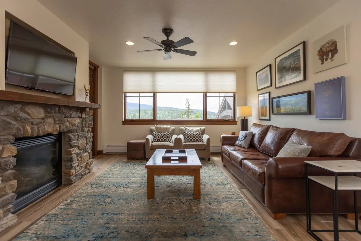This image captures a welcoming living room centered around a striking stone fireplace, flanked by a leather sofa and large windows revealing dramatic mountain views. The space is adorned with decorative artwork and a coffee table, inviting relaxation and social gatherings in a cozy atmosphere.