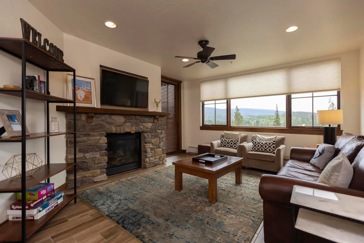 The image shows a warm and inviting living space inside a luxury Durango condo. A plush sofa faces a crackling fireplace, perfect for cozy evenings after a day on the slopes. Large windows offer sweeping sunset views of the surrounding mountains, bathing the room in golden light. A coffee table invites guests to gather for games or relaxing conversations, with every detail curated for comfort and connection.