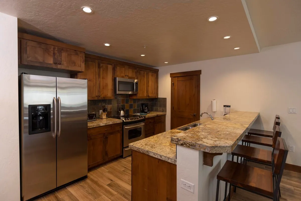 A warmly lit kitchen scene highlighting a stainless steel refrigerator, granite countertops, wooden cabinets, and bar stools. The inviting layout encourages both cooking and casual dining, offering a different angle that complements the primary kitchen image.