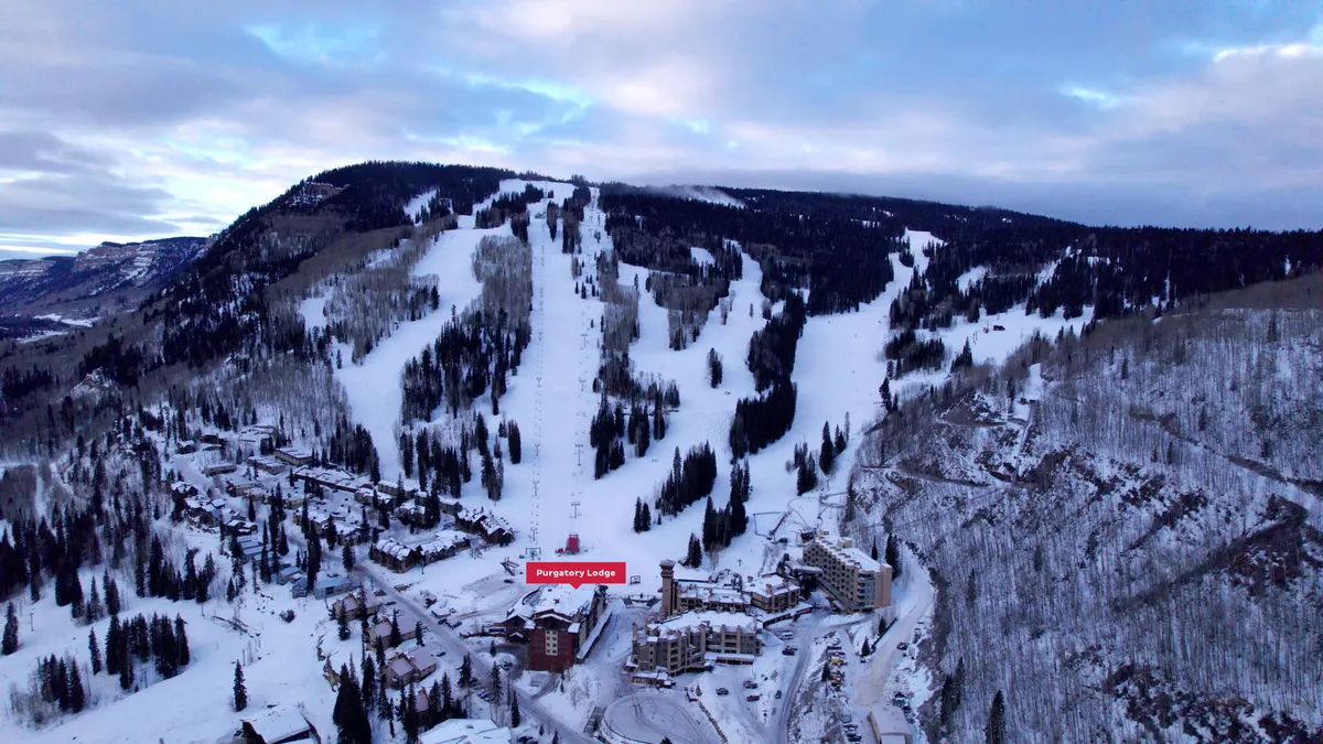 This image captures the snow-covered ski slopes directly outside Purity Lodge, with the lodge nestled among pine trees and the dramatic backdrop of the surrounding mountains. The scene conveys the thrill of ski-in/ski-out access and the inviting atmosphere of returning to a cozy retreat after a day on the slopes.